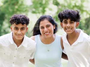 A group of three people wearing white shirts smile together outdoors on a sunny day with greenery in the background.