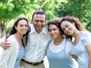 A family group smiles while posing together outdoors against a backdrop of green trees and foliage.