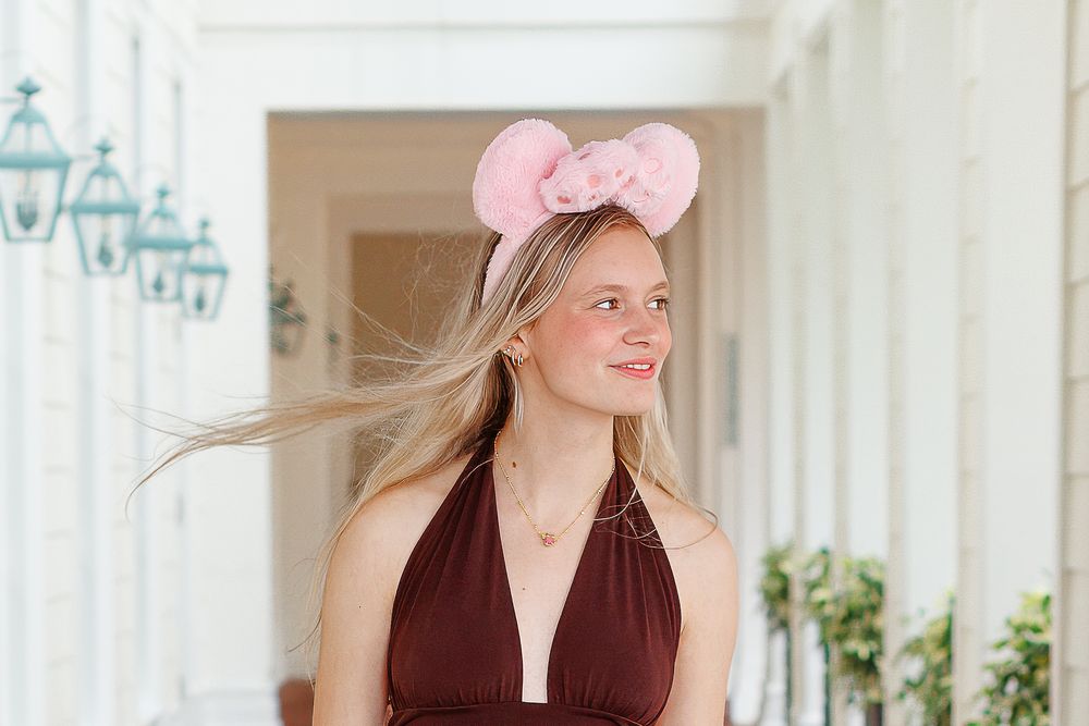 Woman in pink bow headband and brown vest smiles while standing in front of white columns.