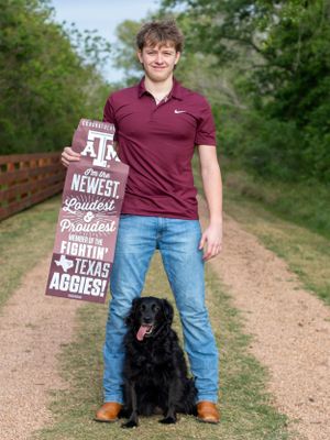 Student poses with a black dog and Texas A&M sign on a wooden bridge pathway.