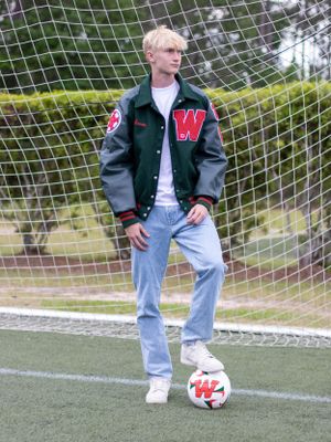 Soccer player in varsity jacket poses with ball in front of goal net.