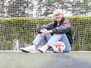 Athletic student in varsity jacket sits casually against soccer goal with ball.