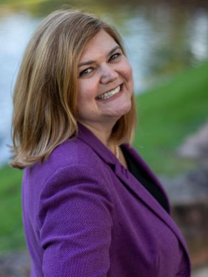 Person in purple blazer poses outdoors with water and greenery in background.