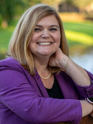 Person in purple blazer smiles warmly while posing outdoors near water.