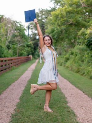 Person in a white dress celebrates graduation by jumping on a dirt path with trees in background.