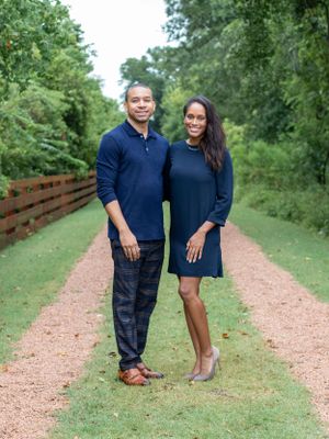 Couple in matching navy outfits poses on gravel path with wooden fence.