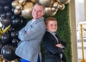 Two people pose back-to-back in front of a balloon arch display.