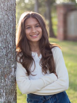 Portrait of a young person in a white sweater leaning against a tree with long dark hair.