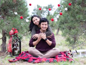 A couple sits on a red blanket surrounded by holiday decorations in a pine forest.