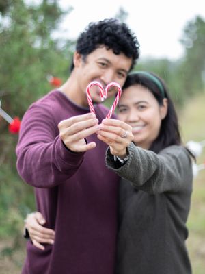 A couple holds candy canes in the shape of a heart with red decorations in the background.
