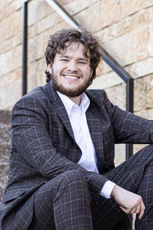 Professional headshot of someone in a dark checkered suit jacket and white dress shirt against a stone wall background.