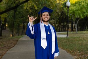 Graduate in blue cap and gown walking and smiling on a tree-lined path during autumn graduation ceremony.
