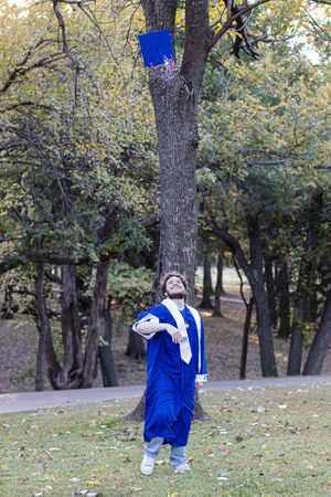 Someone in a blue graduation gown throws their cap in the air during a graduation photo session in a park setting.