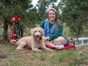 A person sits with a golden doodle dog on a red blanket in a forest setting.
