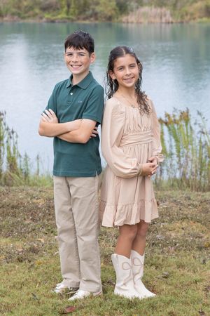 Two children stand together by a lake wearing coordinated outfits on a grassy shore.