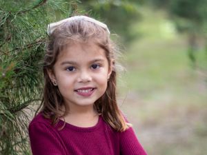 Young child in burgundy sweater smiling next to pine tree outdoors in natural lighting.