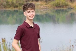 A person in a burgundy polo shirt smiles while standing near a serene lake with natural scenery in the background.