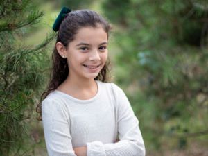 Young person in white sweater poses confidently near evergreen branches.