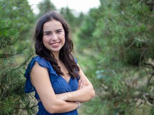 Portrait of person in blue top smiling against pine tree background.