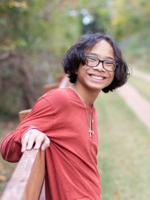 Student in red sweater and glasses leans casually on railing outdoors with natural background.