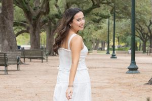 Woman in white dress walking through a park with trees and lampposts on a sunny day.