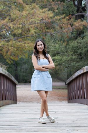 A person in a light blue denim dress and white sneakers stands on a wooden bridge surrounded by autumn foliage.