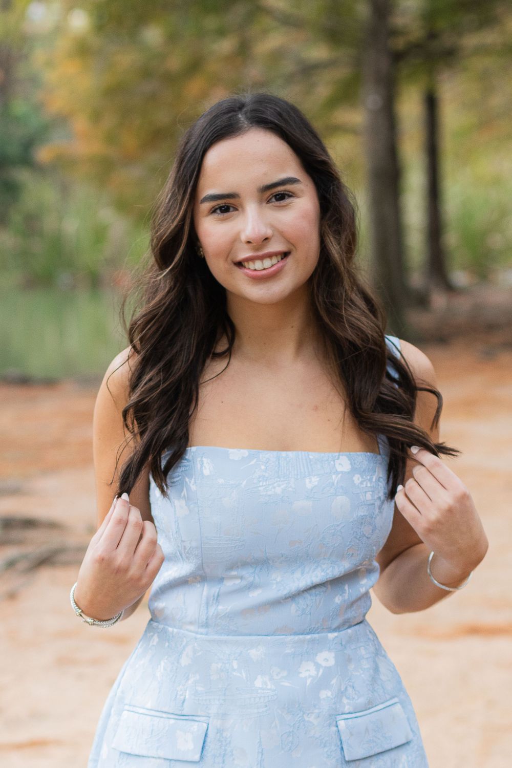 A person in a light blue floral dress poses outdoors in a wooded autumn setting.