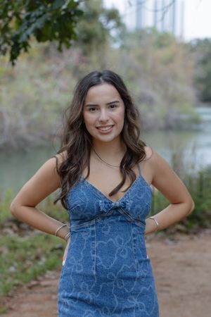 Someone wearing a blue denim dress poses outdoors near a lake with trees in the background.