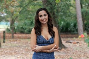 A person in a navy blue dress poses outdoors in a wooded setting with autumn leaves on the ground.