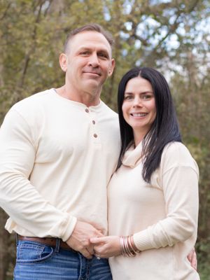 Couple in matching cream sweaters poses together on wooded path.