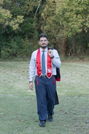 A person in formal attire with a red vest and tie stands outdoors in a grassy field with trees in the background.