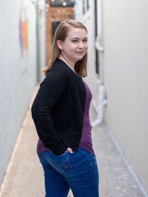 Professional portrait of person in black blazer and purple top standing in hallway.