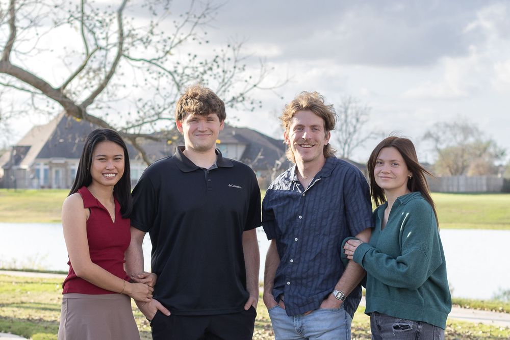 A group of people pose together outdoors in casual clothing against a natural landscape background.