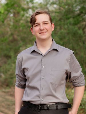 A person wearing a gray button-up shirt and black slacks poses outdoors against a natural green background.