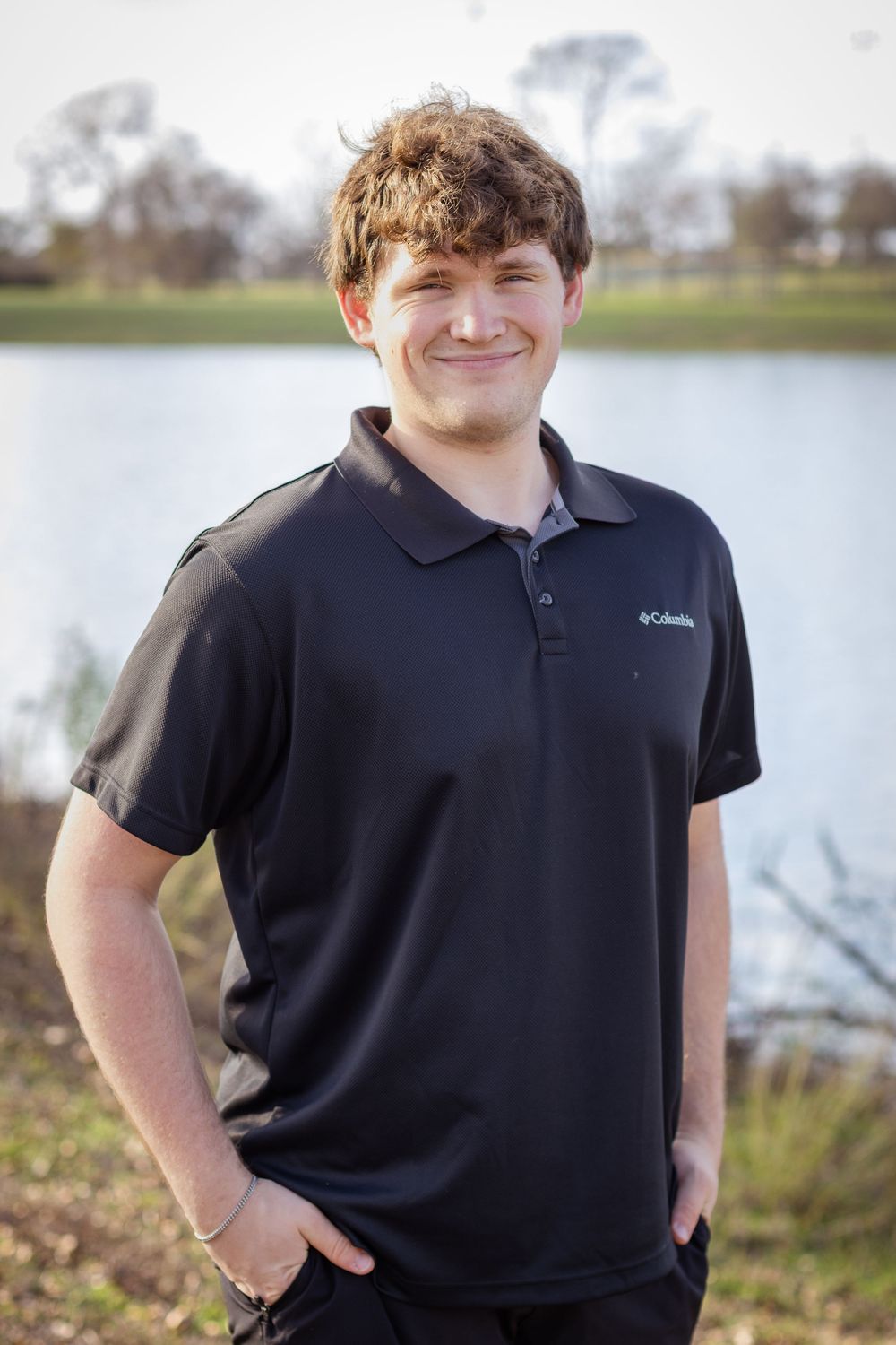 Person in black Columbia polo shirt poses outdoors near a lake on an overcast day.