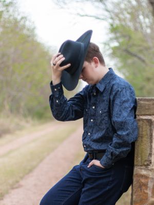 On a dirt path, someone wearing a navy button-down shirt and dark hat leans against a wooden post in a thoughtful pose.