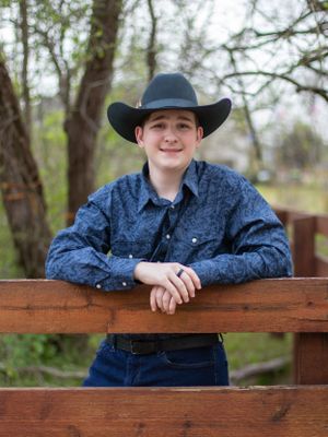 Young person in western attire with cowboy hat and denim shirt leans against wooden fence in outdoor setting.