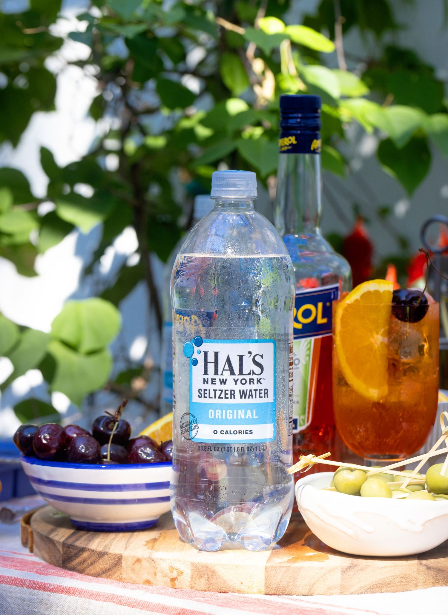 Hal's New York Original Seltzer Water bottle on a wooden board with cherries, olives, and cocktails.