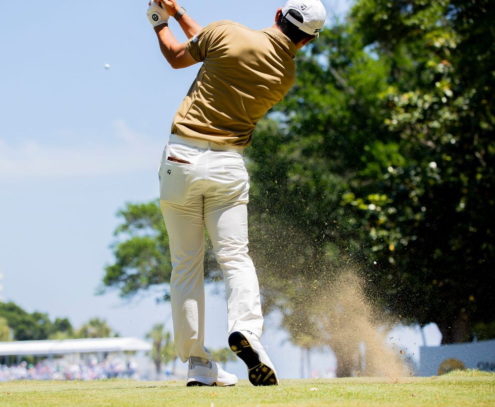 Golfer in tan shirt and white pants swings club on sunny golf course, sending sand flying through the air.