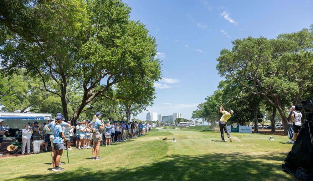 Golfer tees off before a crowd of spectators on a sunny golf course lined with large green trees.