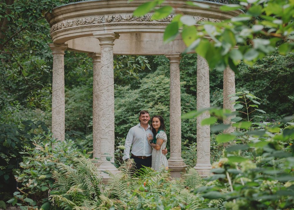 Smiling couple standing together under an ornate stone gazebo surrounded by lush green foliage.