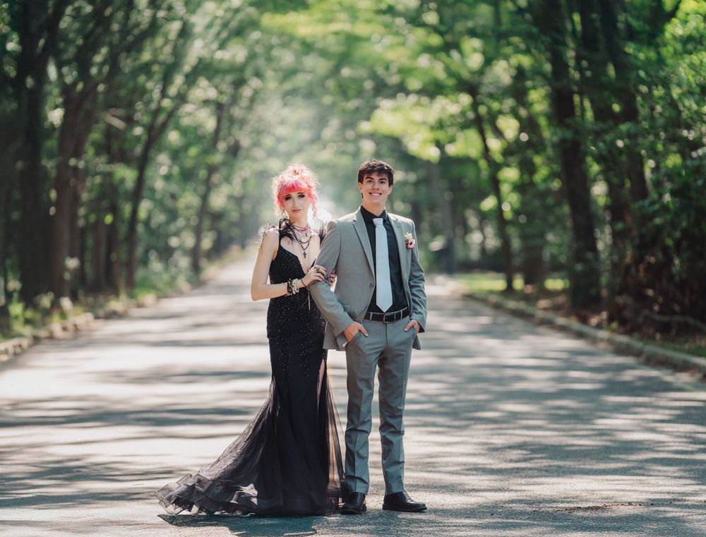 Couple in formal attire poses on tree-lined road for prom portraits.