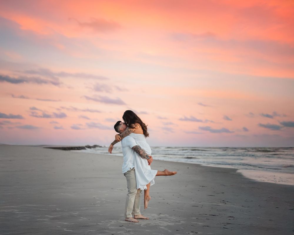 Man lifting woman in white dress on a beach during a vibrant pink sunset by the ocean.