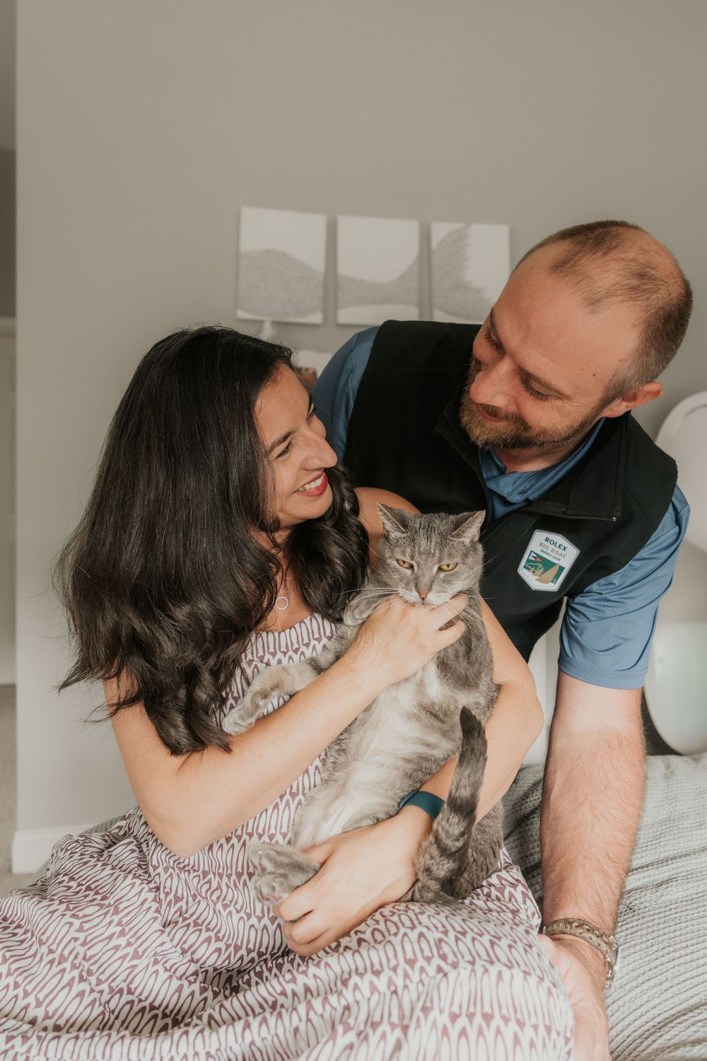Smiling couple sitting on a bed together holding a gray cat in a cozy indoor setting.
