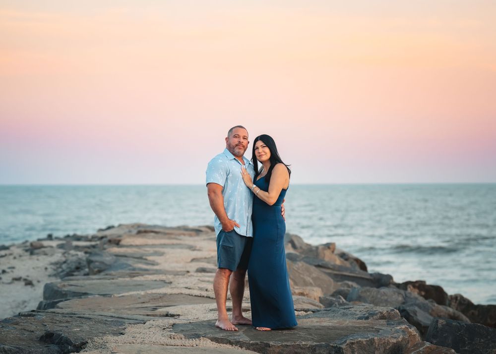 Couple embraces on rocky jetty at sunset with ocean background.