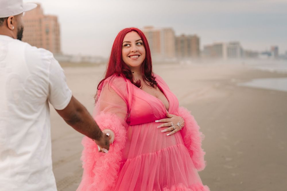 Pregnant woman in pink tulle maternity gown holds partner's hand on beach.