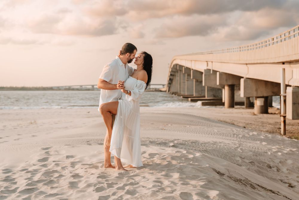 Couple sharing a romantic moment on a sandy beach near a pier during a soft sunset.
