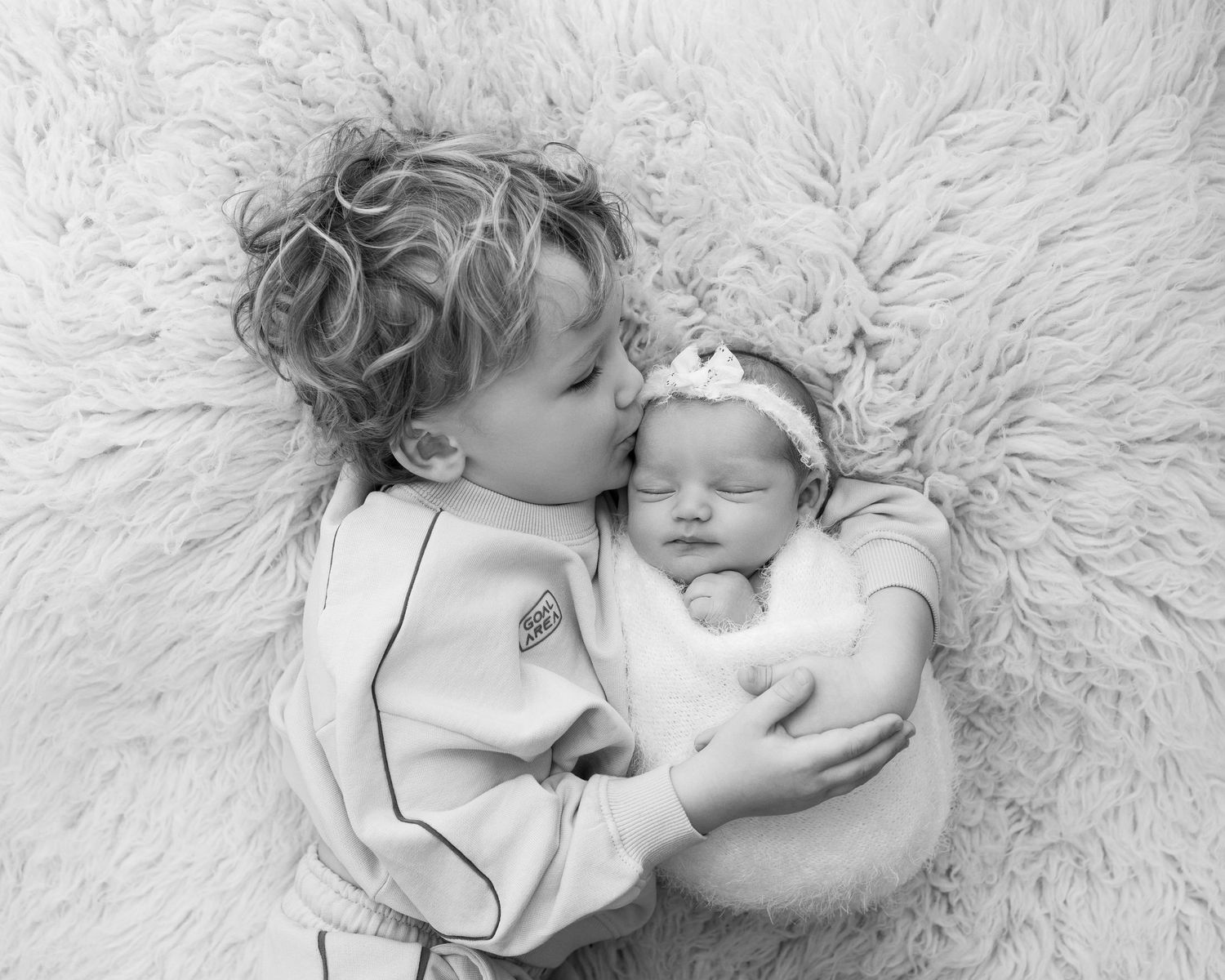 A black and white photograph of two young siblings cuddling on a plush furry rug.