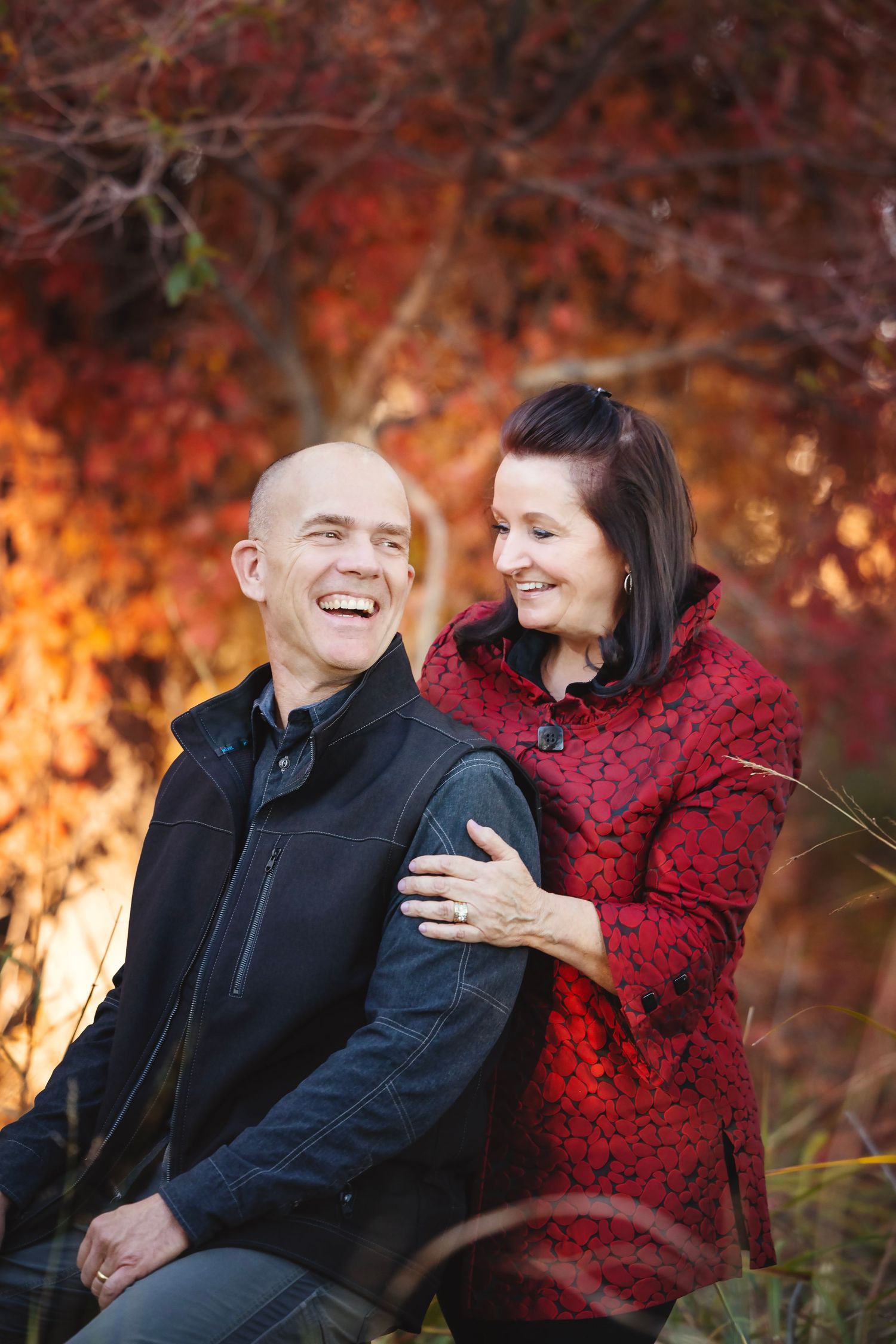Smiling couple posing outdoors in front of vibrant red and orange autumn foliage.