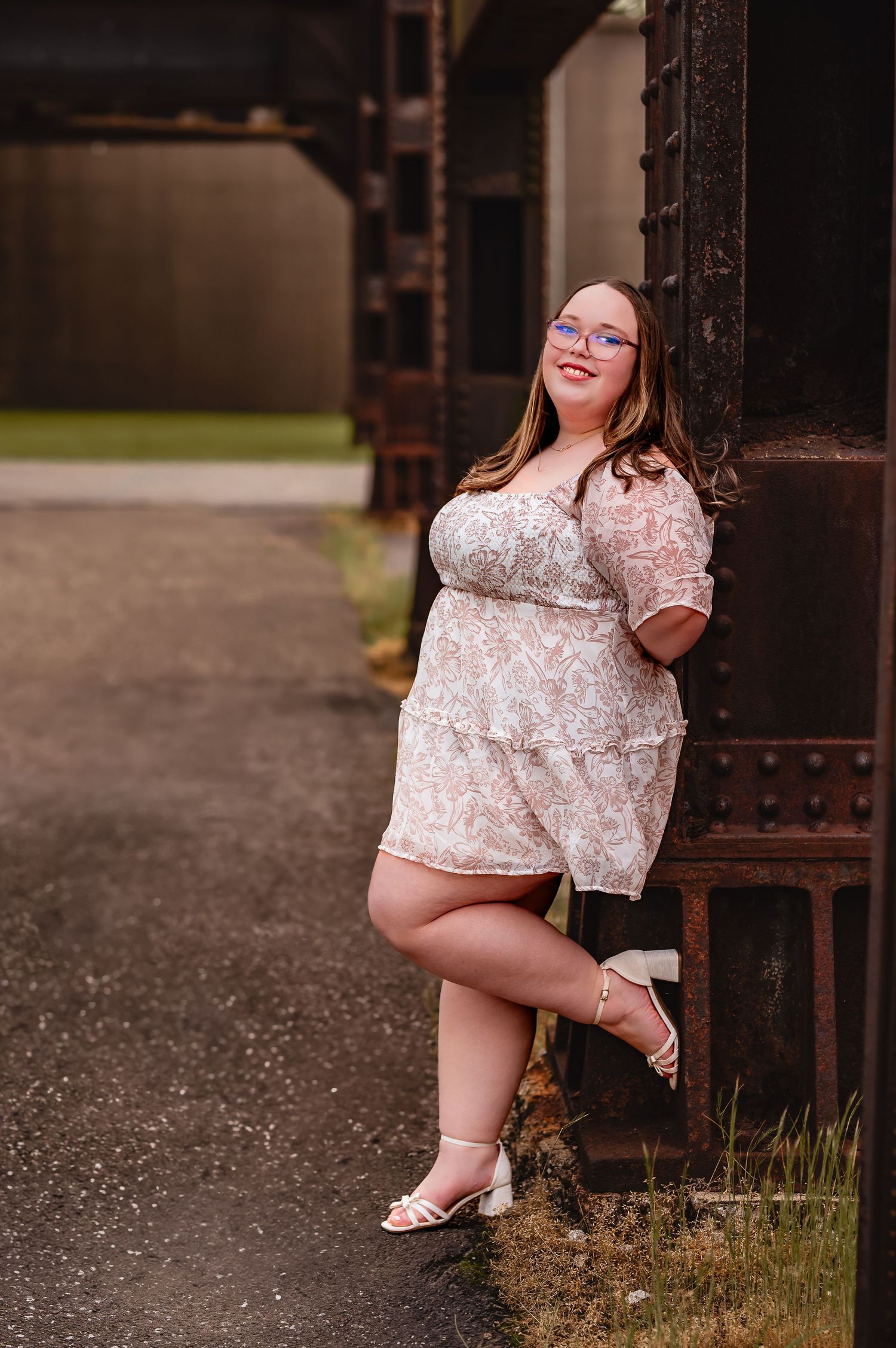 A woman in a lace dress poses confidently against an industrial metal structure.